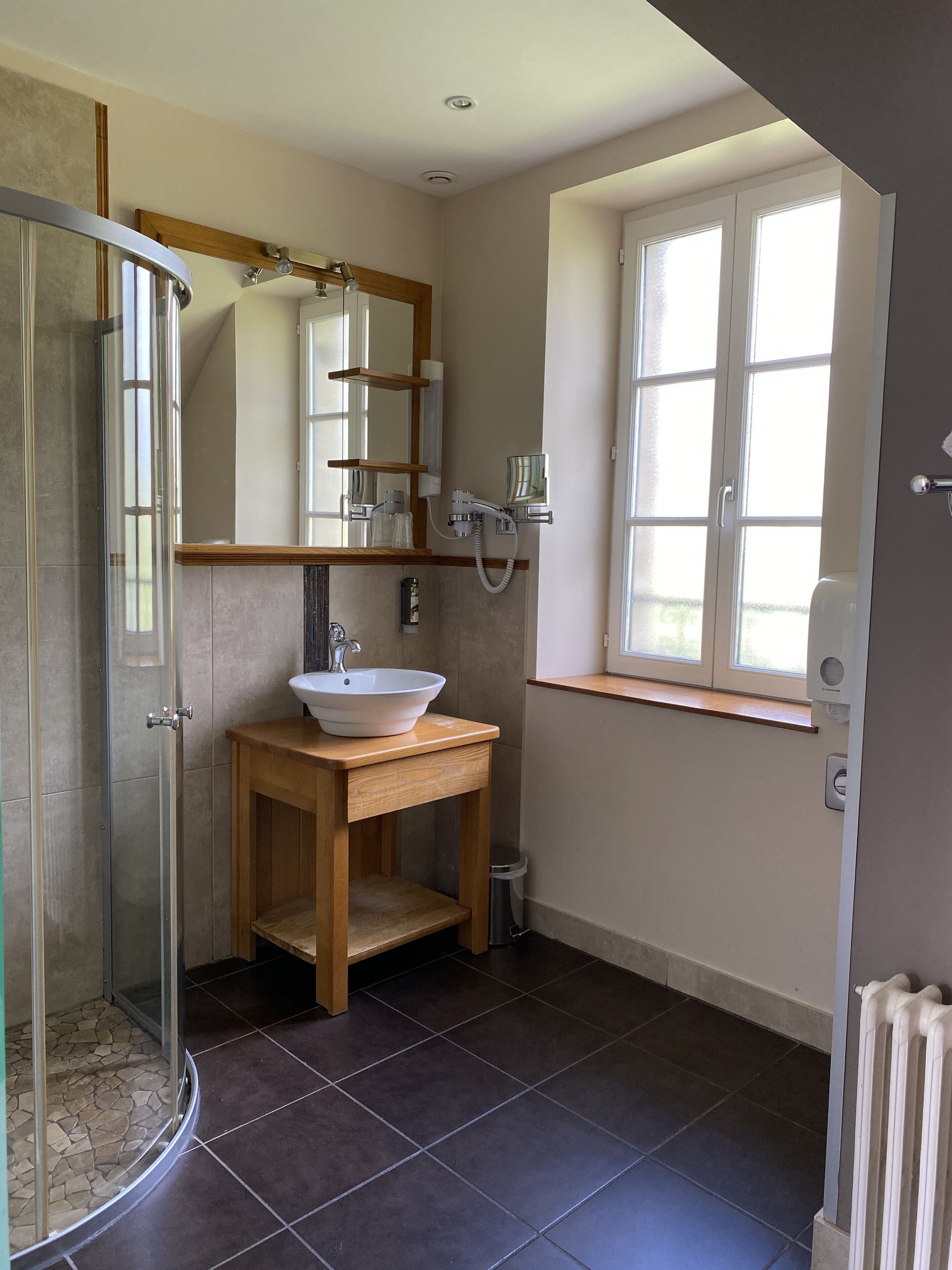 Un baño con cabina de ducha de vidrio, tocador de madera con lavamanos blanco, espejo sobre el lavamanos y ventana con marcos blancos.