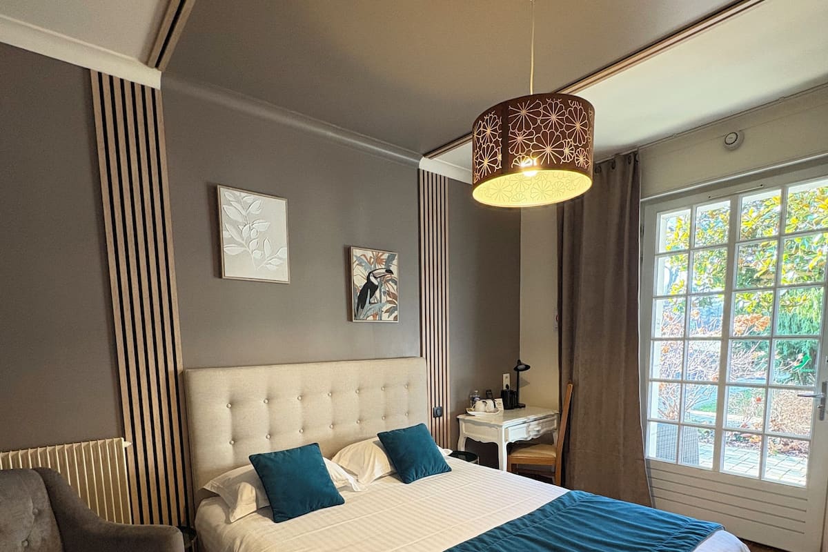 A modern bedroom with a large bed, a grey armchair, a bedside table, and a window with a view of trees.