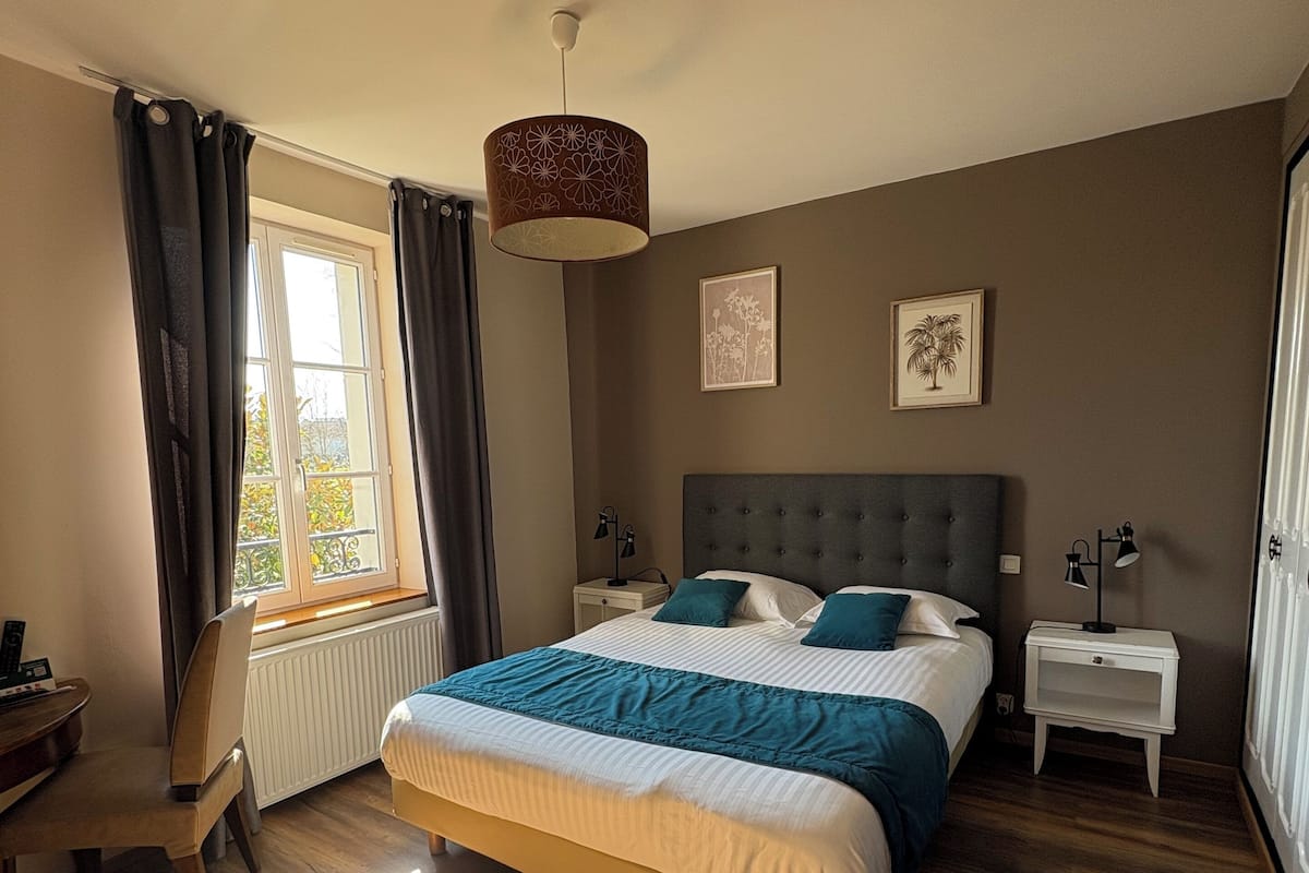 A bedroom with a large bed, a desk, and a chair. There is a window with curtains, a radiator, and a ceiling light fixture.