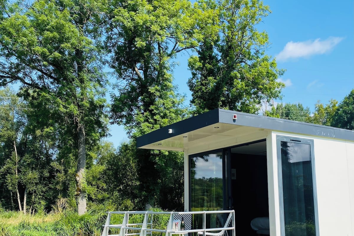 A modern houseboat with a glass door, a small deck, and a view of the surrounding greenery.