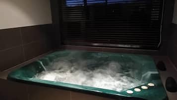 Una vasca idromassaggio con interno verde, circondata da piastrelle scure, e una finestra con persiane sullo sfondo.