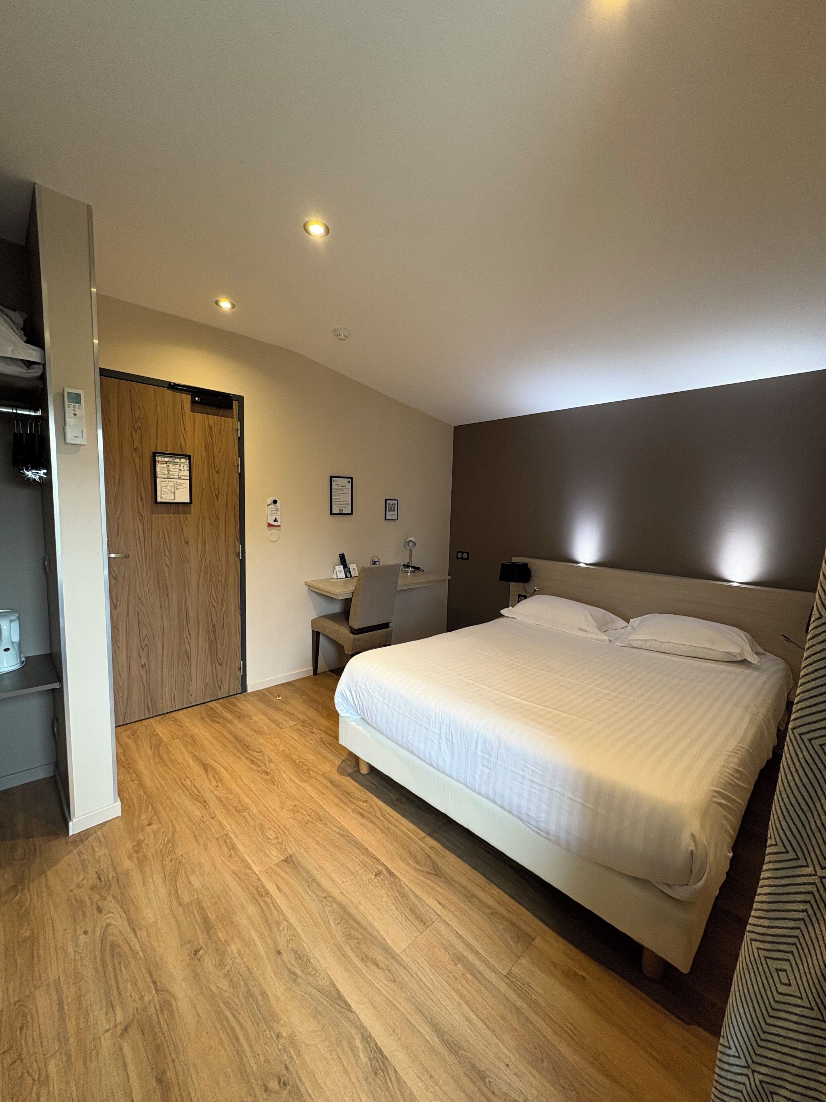 Una habitación de hotel con piso de madera, una cama con ropa blanca, un escritorio con una silla, un armario de madera y una puerta con un letrero.
