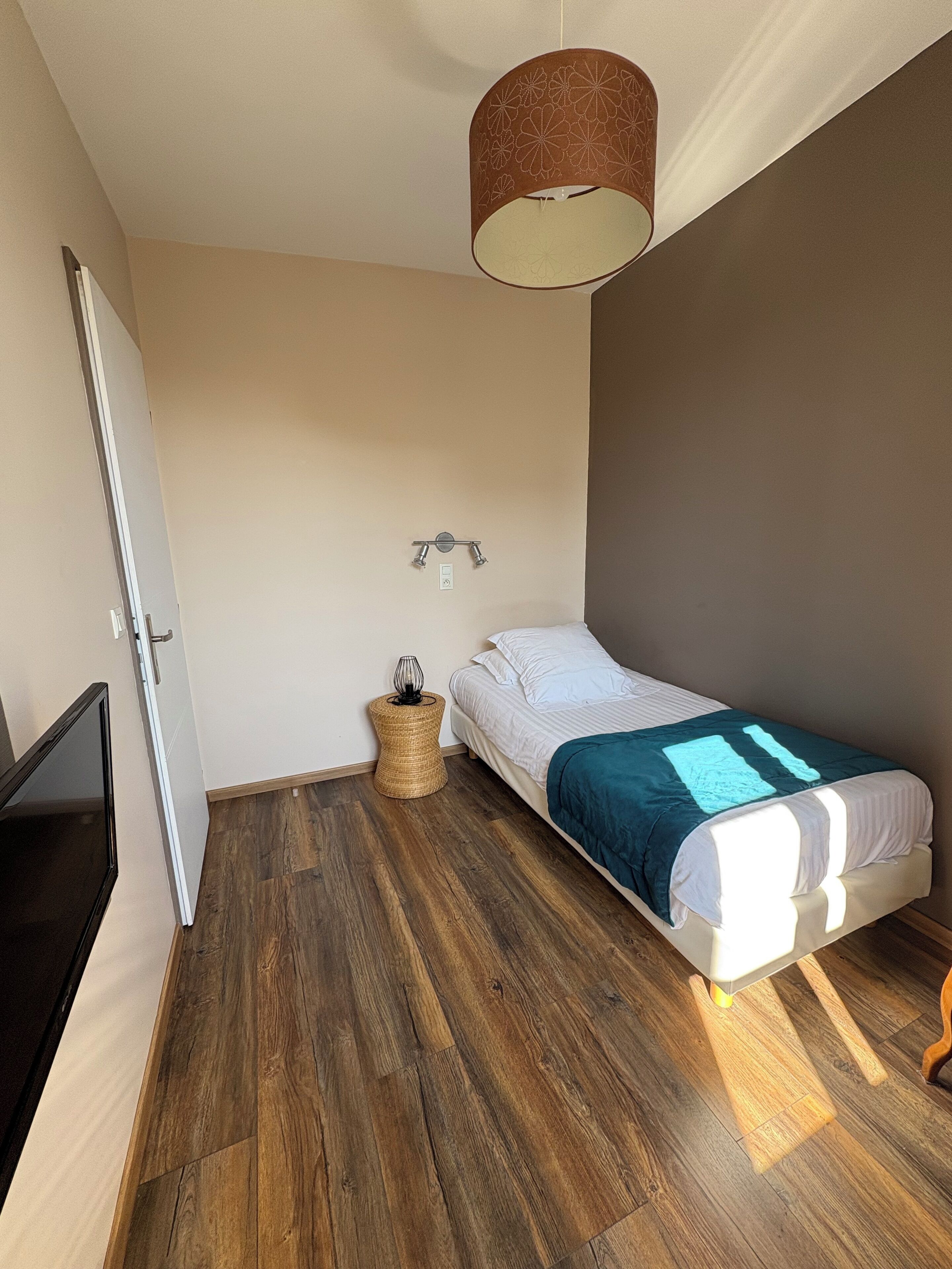 A small, modern bedroom with a single bed, a TV, and a ceiling light fixture.