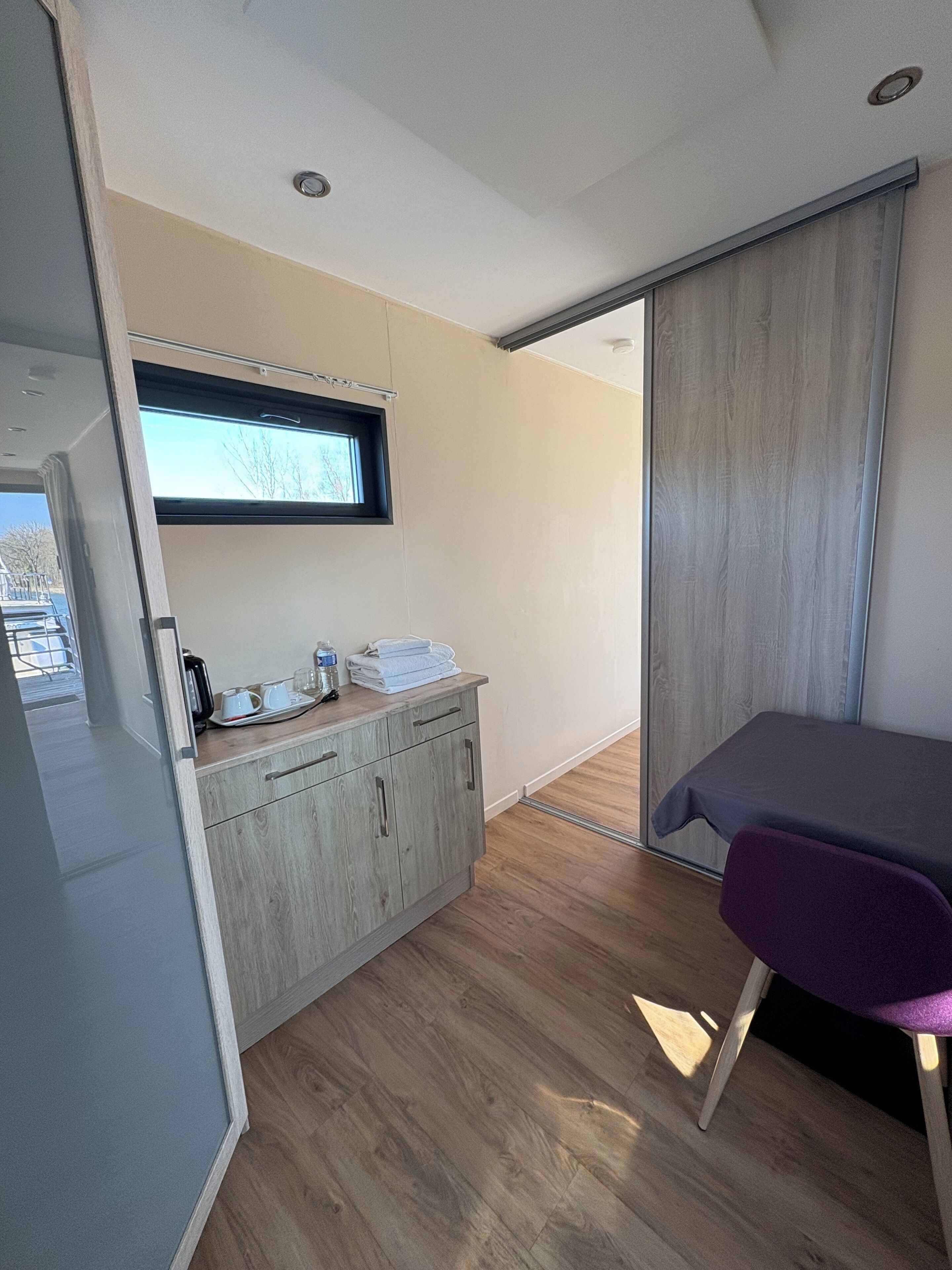 Een moderne, compacte kamer met een kitchenette, een klein raam, een schuifdeur en een paarse stoel.