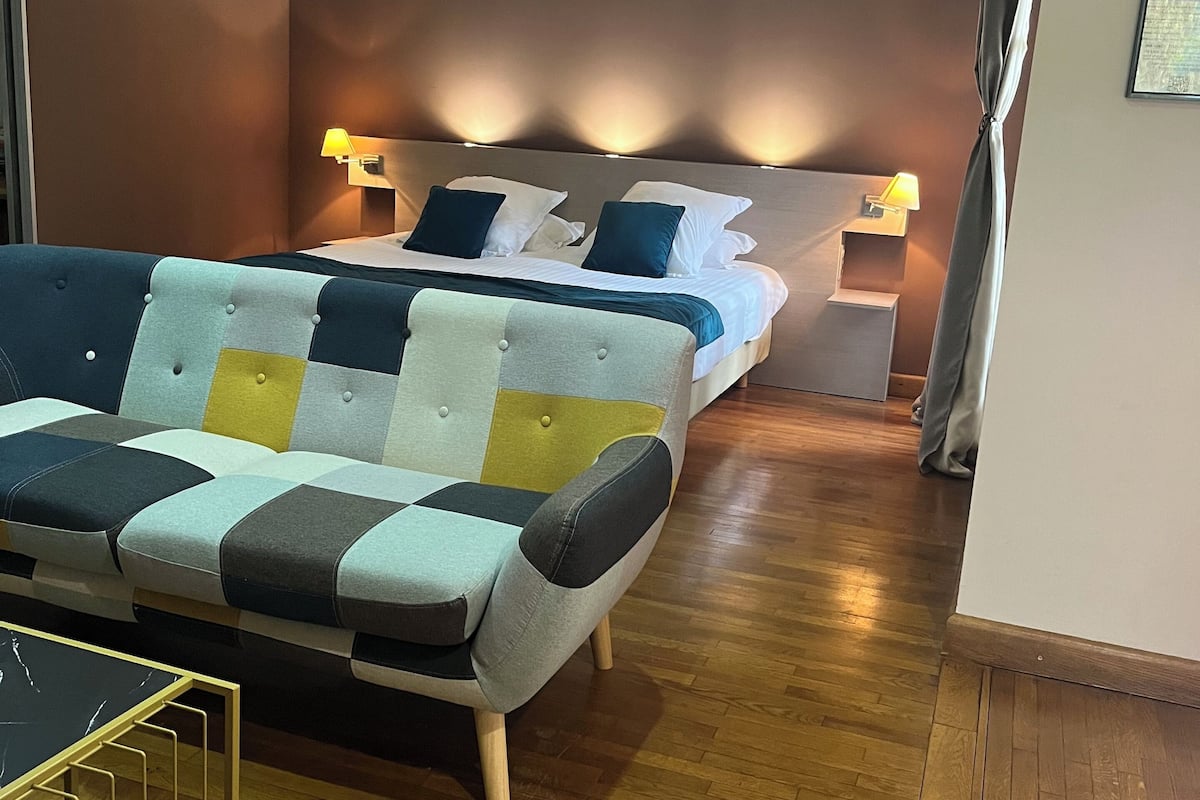 A hotel room with a sofa, a bed with two pillows, a nightstand with a lamp, and a wooden floor.