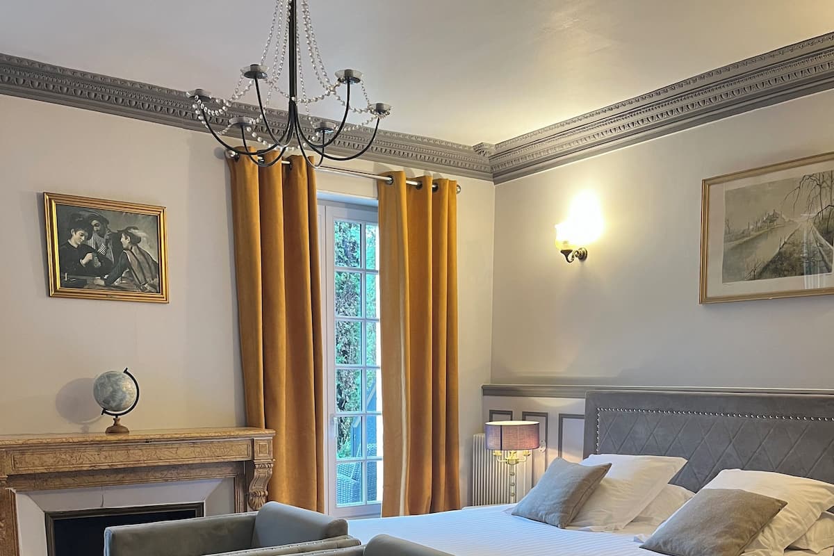 A bedroom with a large bed, two armchairs, a fireplace, a chandelier, and a window with curtains.