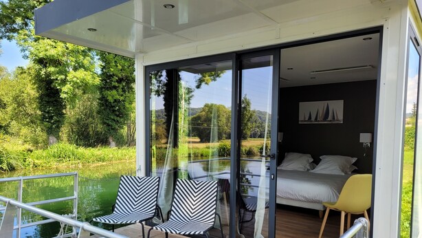 A modern room with a bed, a chair, and a table, connected to a balcony with a view of a lake and greenery. - Hôtel de la Vinaigrerie (Joinville)
