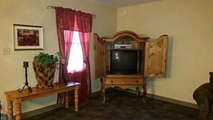 A living room with a wooden TV stand, a bench, a potted plant, and a window with red curtains. - Taneycomo Lakefront Resort (Branson)
