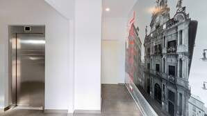 Un ascensor moderno, un pasillo con suelo de baldosas y un mural de un edificio histórico.