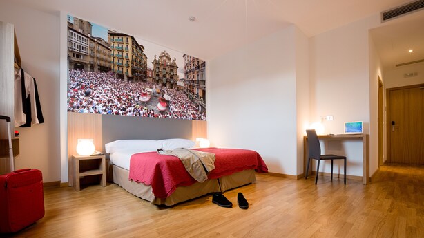 Habitación de hotel con cama, escritorio, silla, maleta roja y un gran mural en la pared que representa un evento concurrido.