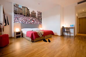 A hotel room with a bed, a desk, a chair, a red suitcase, and a large mural of a crowded event on the wall. - IRAIPE PAMPLONA HOSTAL (Ansoain)