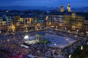 Una plaza con una fuente central rodeada de edificios, una multitud de personas y una torre destacada al fondo.