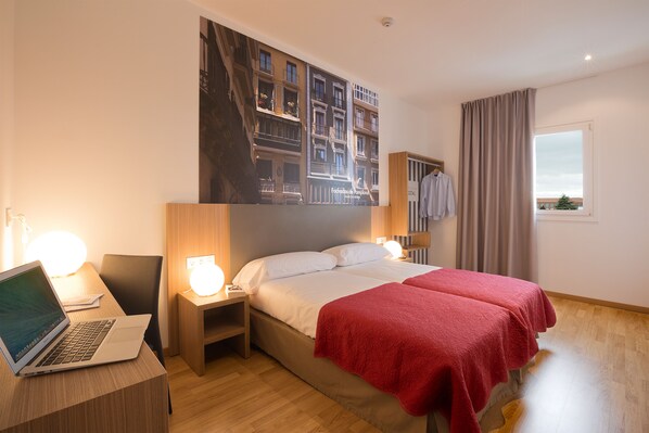 A hotel room with a bed, a desk with a laptop, a nightstand with a lamp, and a large window with curtains. - IRAIPE PAMPLONA HOSTAL (Ansoain)
