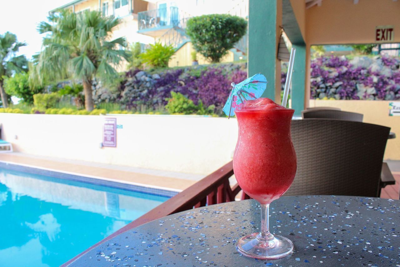 A poolside table with a drink and a view of a pool and greenery.