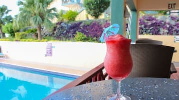 A poolside table with a drink and a view of a pool and greenery.