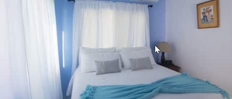 Een slaapkamer met een bed, witte gordijnen, een plafondventilator, een airconditioning, een nachtkastje met een lamp en een lijst aan de muur.
