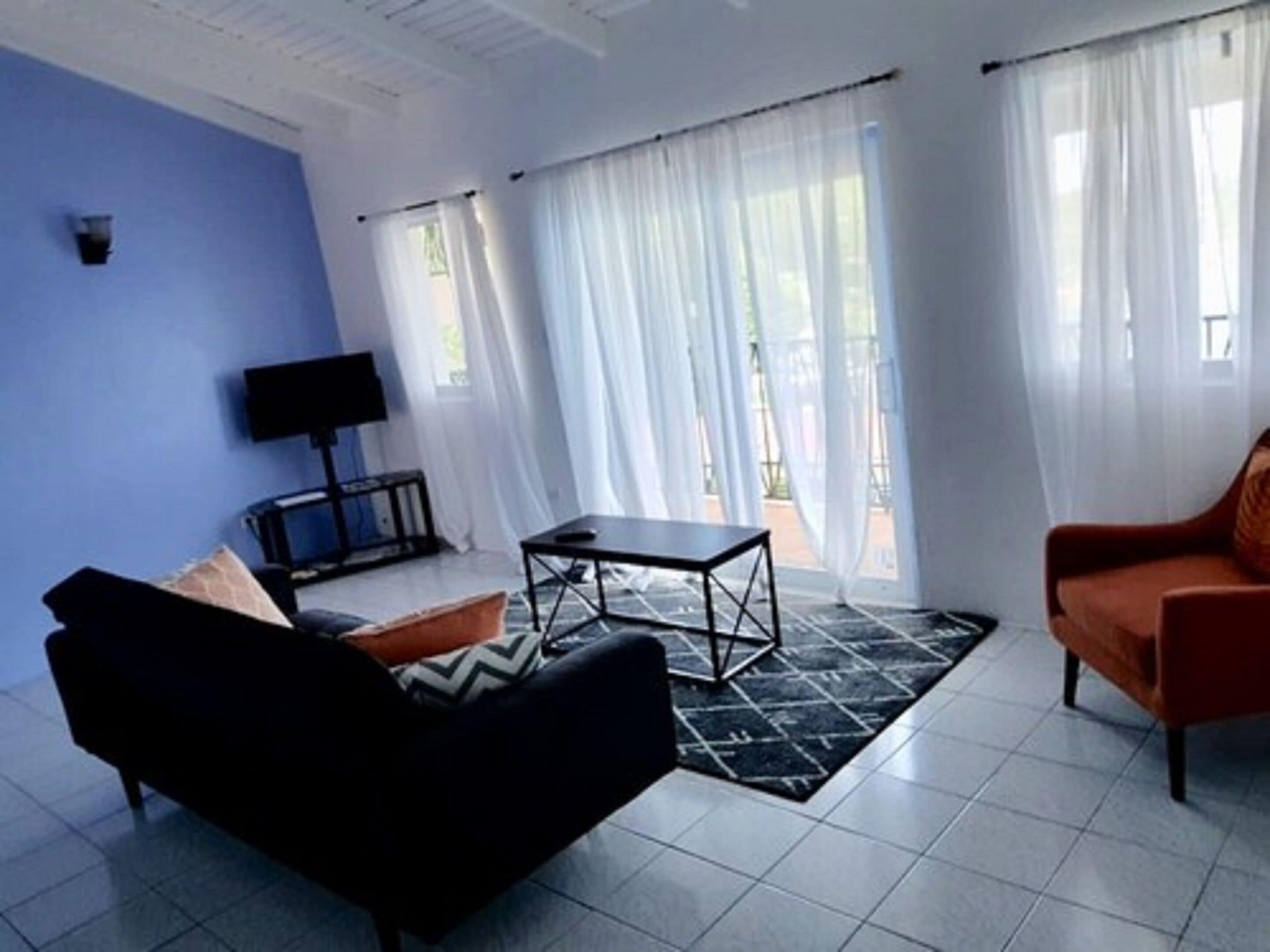 A living room with a black sofa, a small table, and a red armchair. There is a TV on a stand and a window with white curtains.