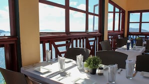 A dining area with tables set for two, large windows with a view of the sea, and a potted plant on the table. - The Heritage Inn (Cane Garden Bay)