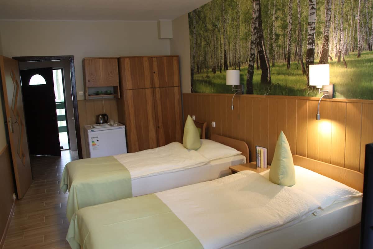 A hotel room with two beds, a wooden wardrobe, a desk with a lamp, and a painting of a forest on the wall.