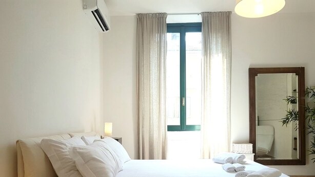 Quarto de hotel com cama, mesa de cabeceira com luminária, janela com cortinas, espelho e planta.