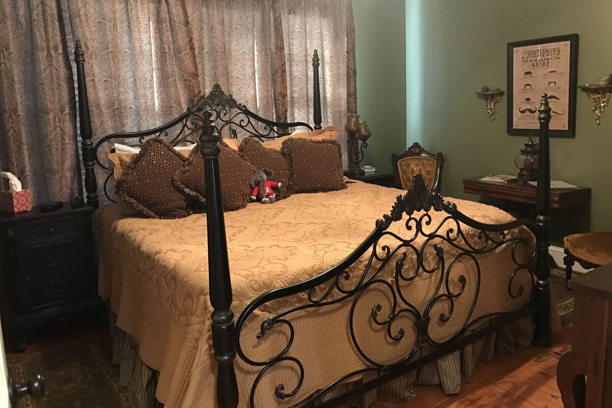 Una cama con dosel con estructura metálica oscura, una colcha estampada y varios cojines decorativos. Hay una mesita de noche con una lámpara y un libro enmarcado en la pared.