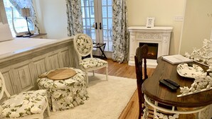 A dining area with a wooden table, floral upholstered chairs, a white fireplace, and a view of the outside through large windows. - Bama Bed & Breakfast Campus (Tuscaloosa)