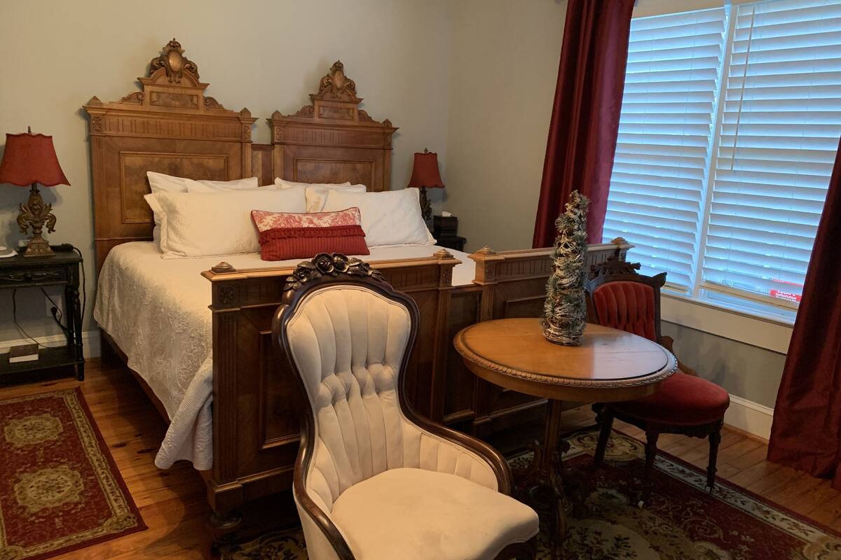 Un dormitorio con una cama de madera, dos mesitas de noche con lámparas, una mesa redonda de madera, un sillón blanco tapizado y una ventana con persianas.