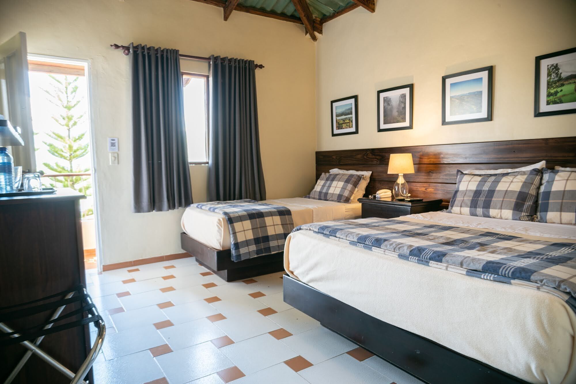 Quarto de hotel com duas camas, uma escrivaninha e uma janela com cortinas. Quadros emoldurados decoram a parede e há um abajur na mesa de cabeceira.