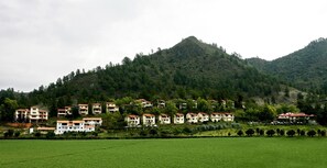 A residential area with multi-story buildings, a green field in the foreground, and a forested hill in the background. - Altocerro Villas Hotel & Camping (Constanza)