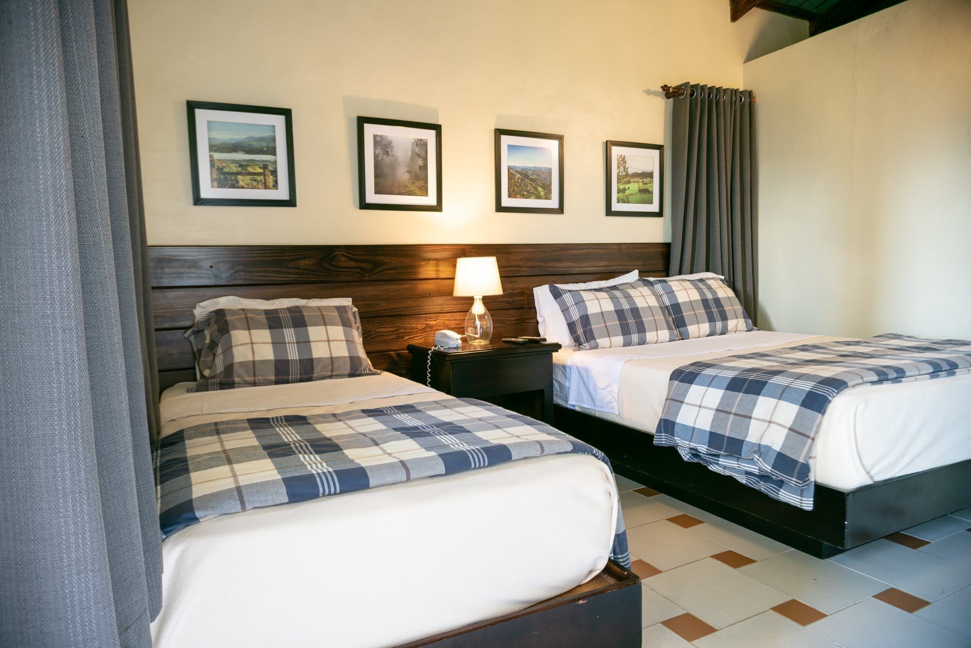 Habitación de hotel con dos camas, cabecero de madera, mesita de noche con lámpara y tres cuadros enmarcados en la pared.