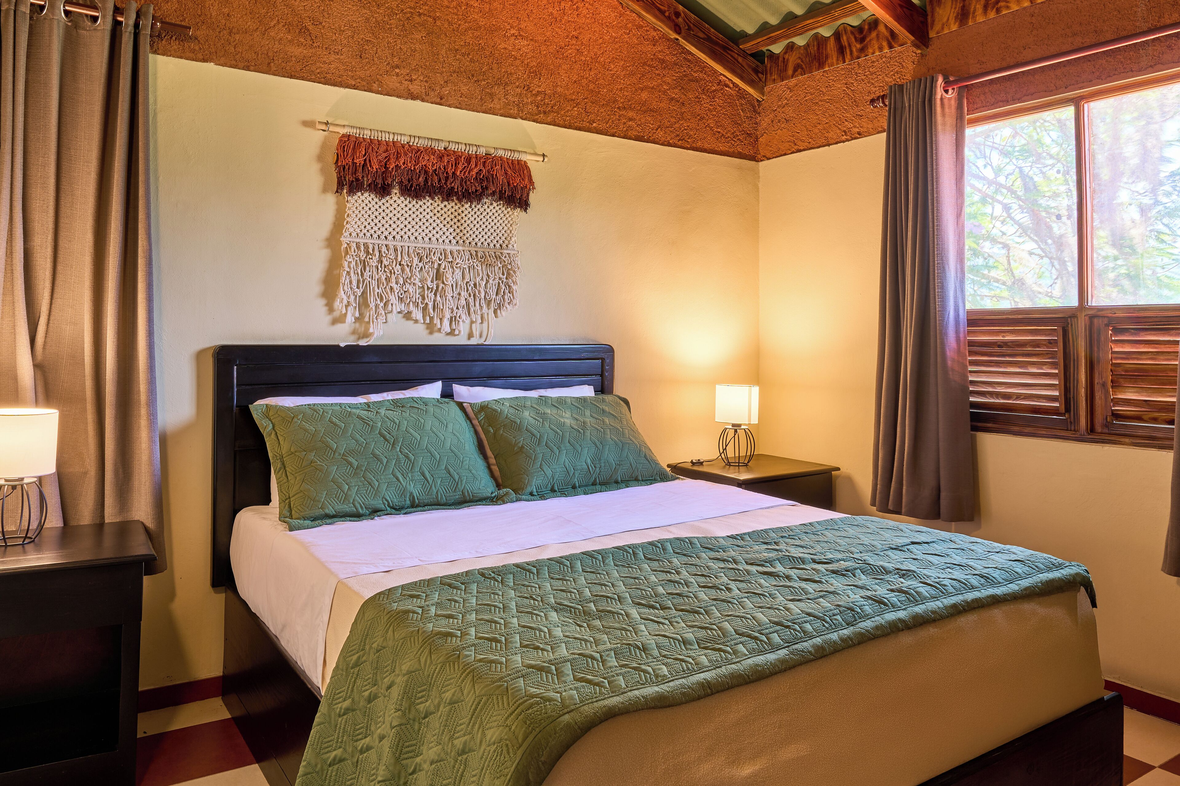 A neatly made bed with a patterned bedspread and pillows, a bedside lamp, and a macrame wall hanging.