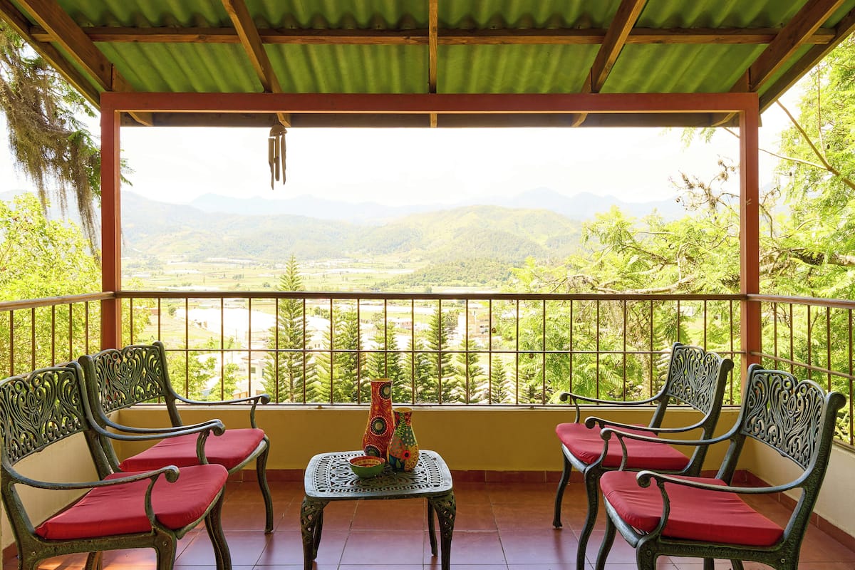 Een balkon met twee stoelen en een tafel, met uitzicht op een schilderachtig landschap met bergen en begroeiing.
