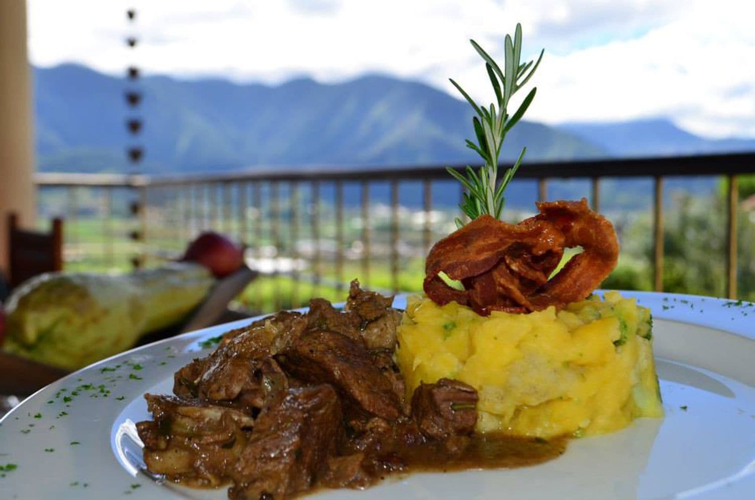 Plato de comida con carne y puré de papas, adornado con romero, en un balcón con vista a la montaña.