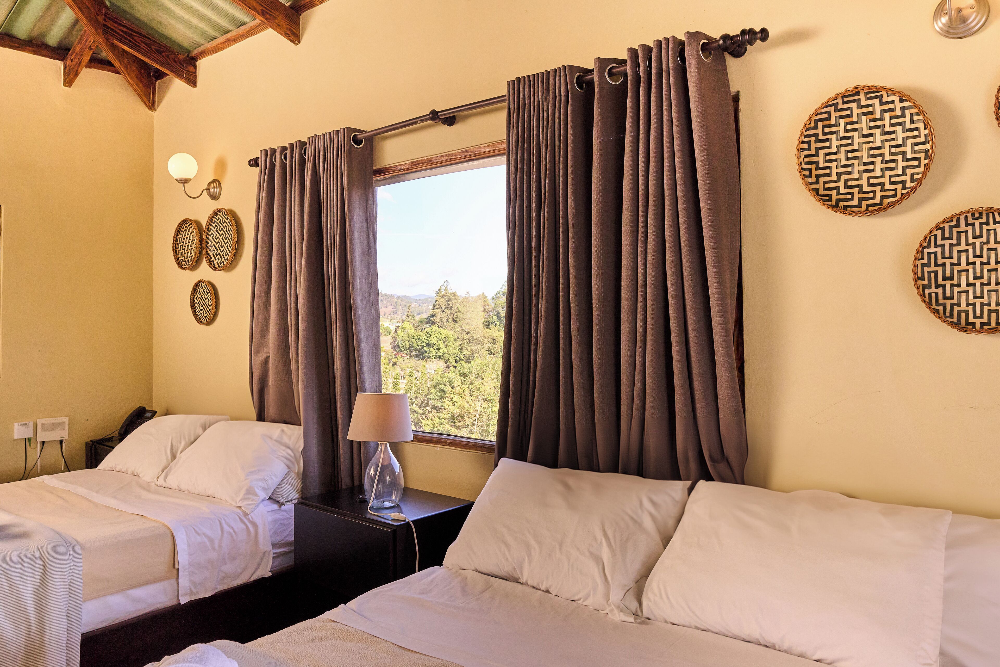 Quarto de hotel com duas camas, uma mesa de cabeceira com abajur, uma janela com cortinas e painéis decorativos na parede.