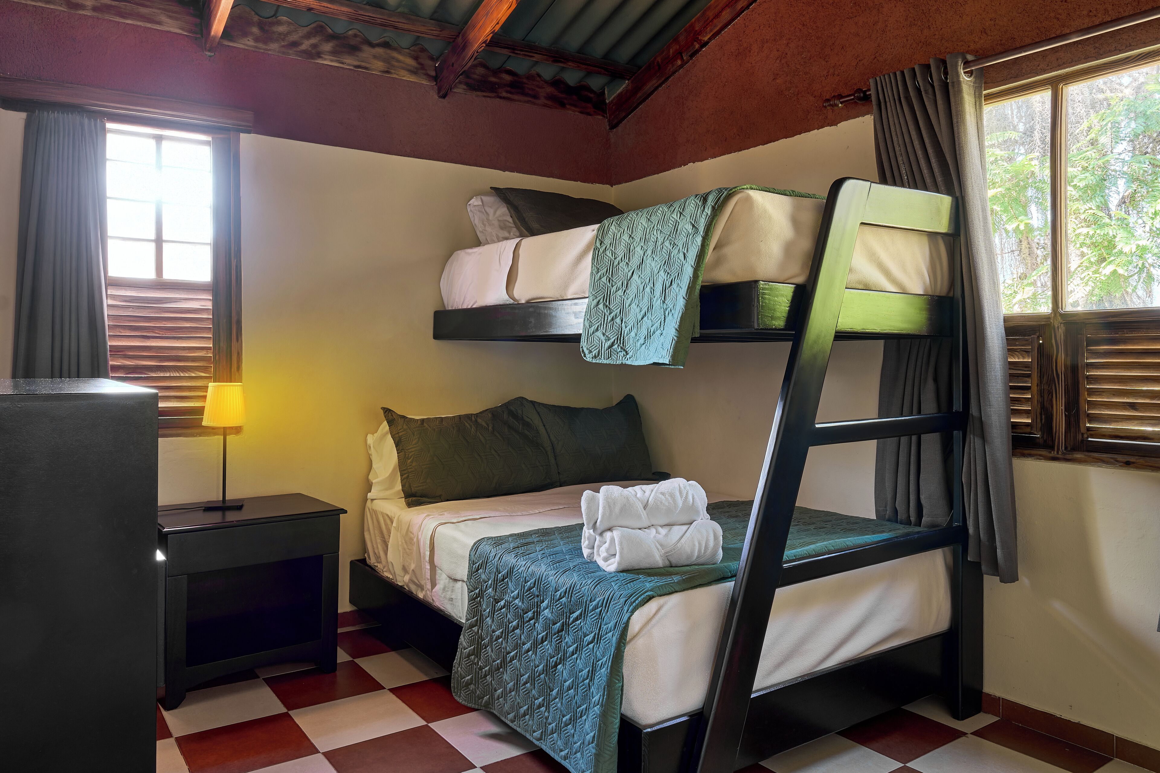 A bunk bed room with a checkered floor, a bedside table with a lamp, and a window with curtains.