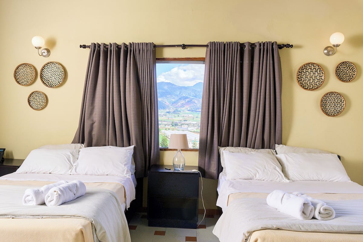 Hotelkamer met twee bedden, een nachtkastje, een lamp en uitzicht op een bergachtig landschap door het raam.