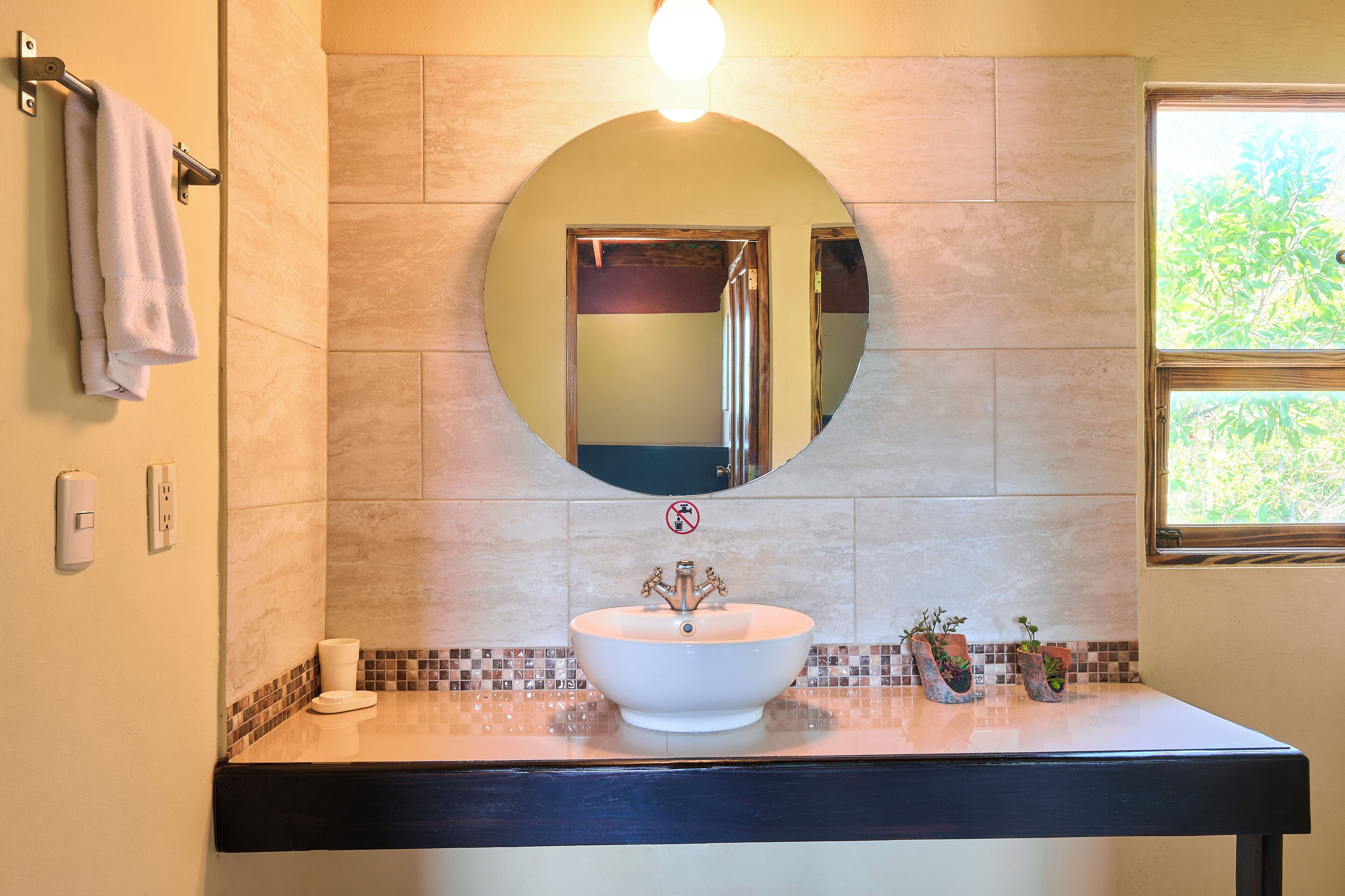 Un baño con un espejo redondo, un lavabo blanco y una ventana con vistas a la vegetación.