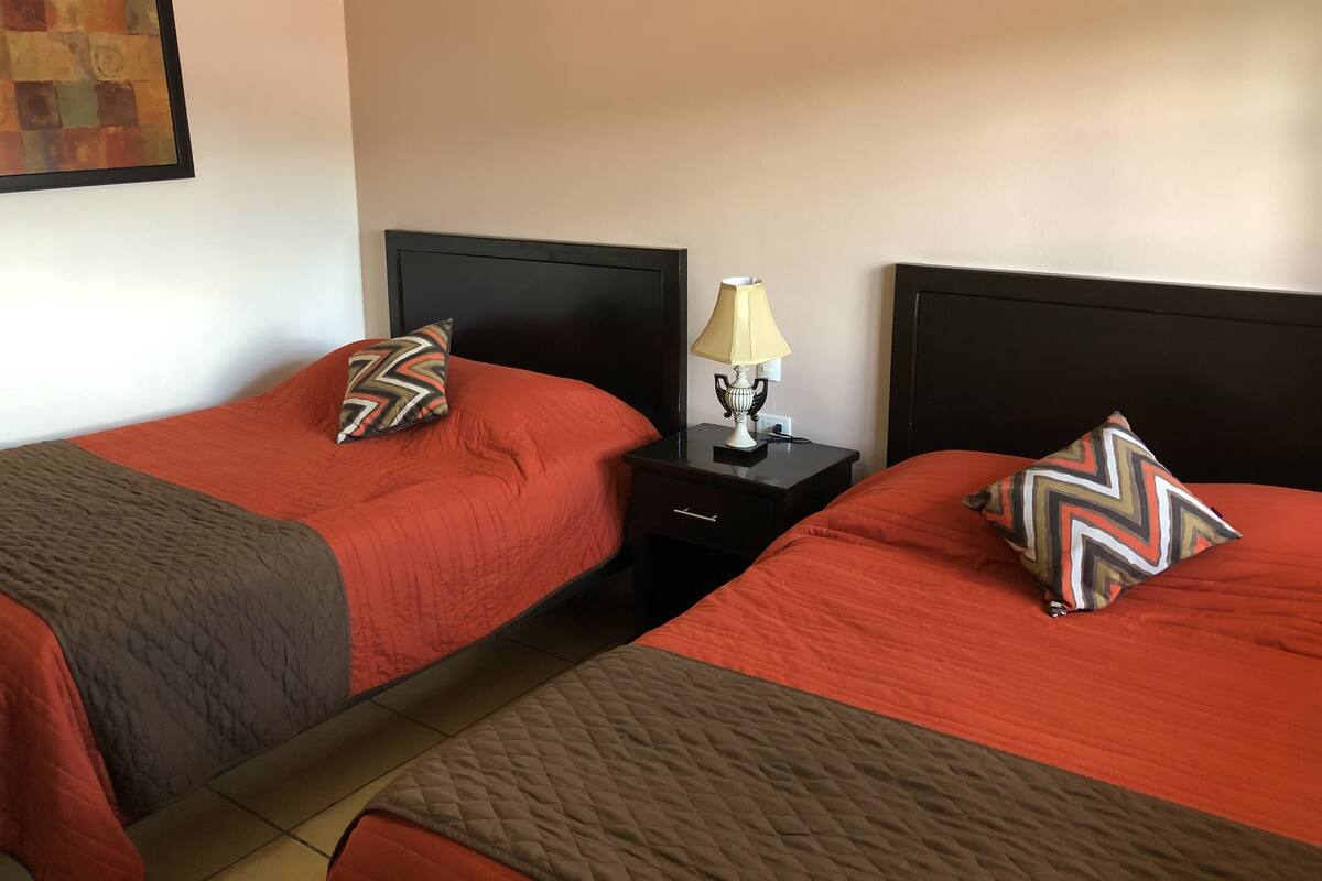 Dos camas individuales con ropa de cama roja, cabeceros negros y almohadas decorativas. Entre las camas, una mesita de noche con lámpara.