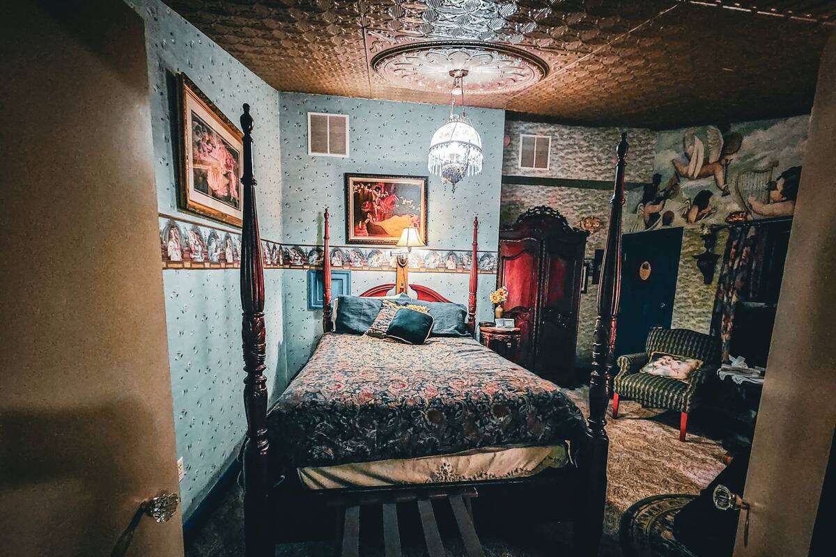 A bedroom with a four-poster bed, a chandelier, a wardrobe, and a mural on the wall.