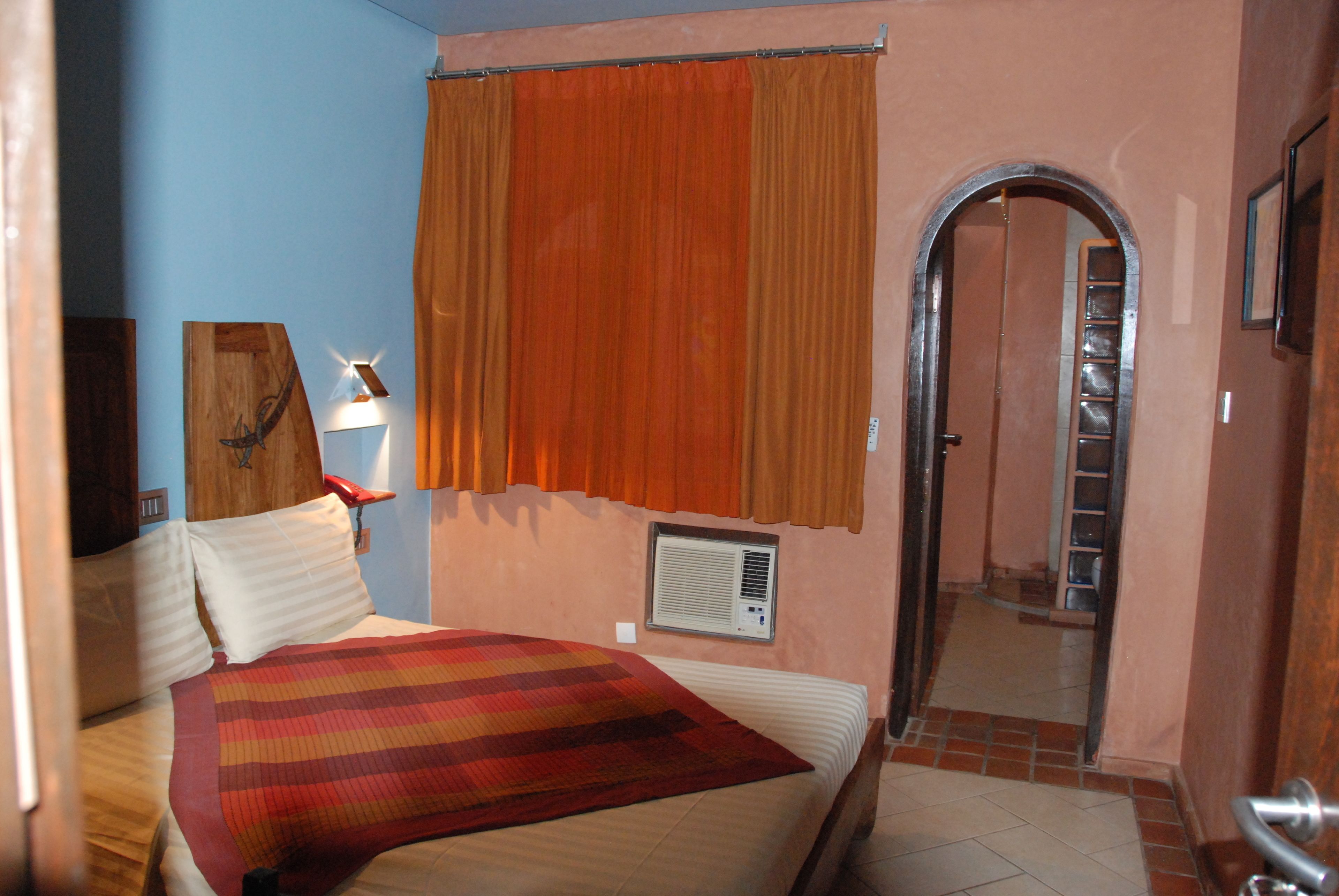 A bedroom with a bed, a window with curtains, a wall-mounted air conditioner, and a door leading to another room.
