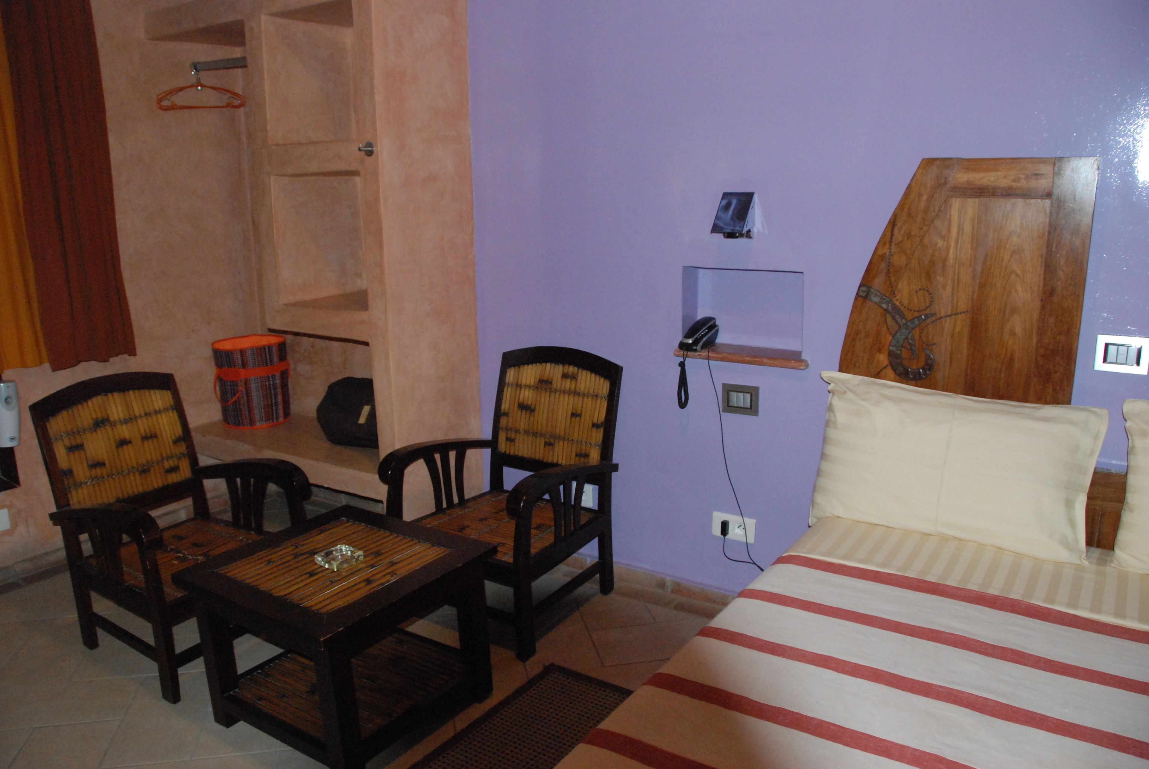 A room with a bed, two wooden chairs, a small table, a hanging coat rack, and a shelf with a telephone and a picture frame.