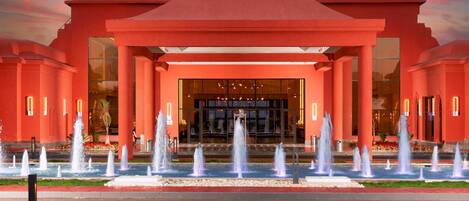 Fachada roja de un hotel, con varias fuentes y una amplia puerta de vidrio.