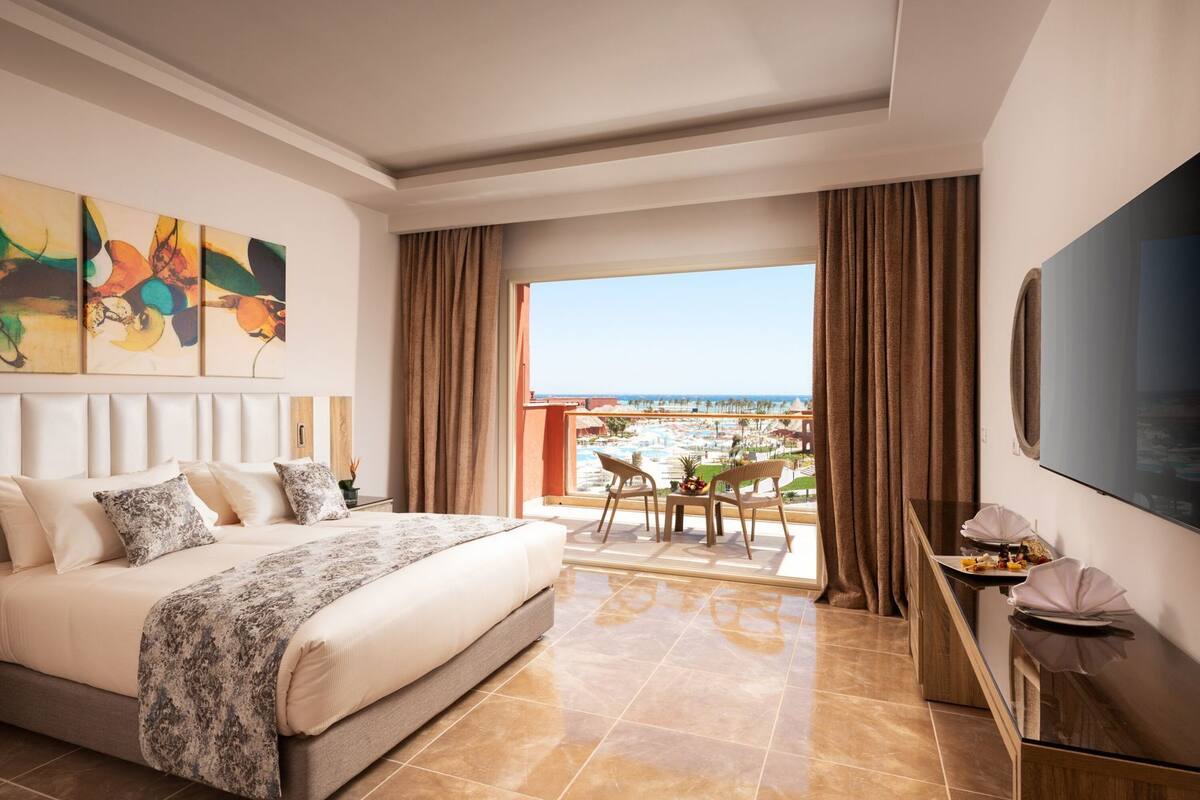 A hotel room with a bed, a TV, a balcony with a view of the beach, and a dining area.