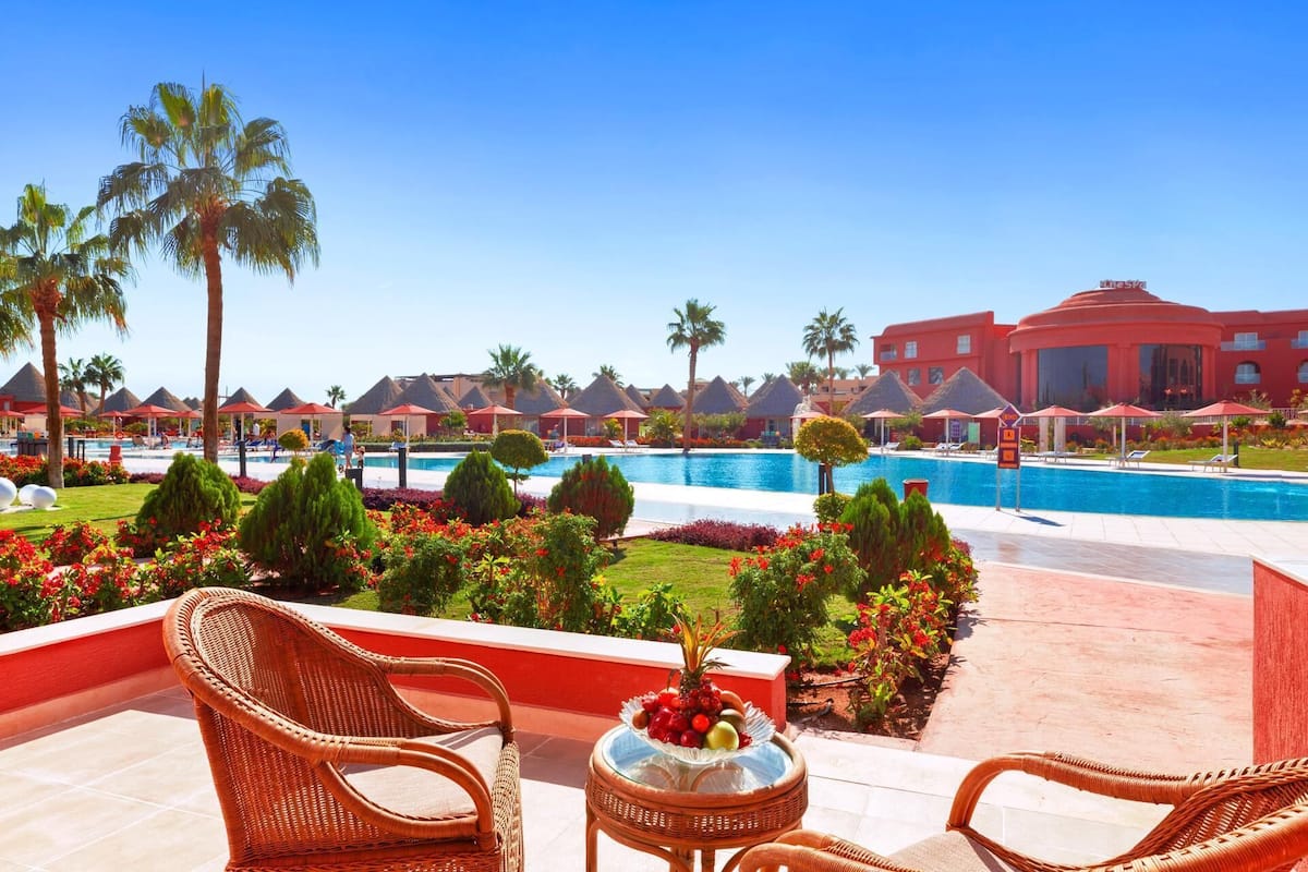 A resort pool area with lounge chairs, a dining table with fruit, and a view of the pool and resort buildings.