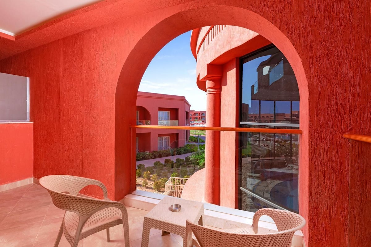 A balcony with two wicker chairs and a small table, overlooking a courtyard with buildings in the background.