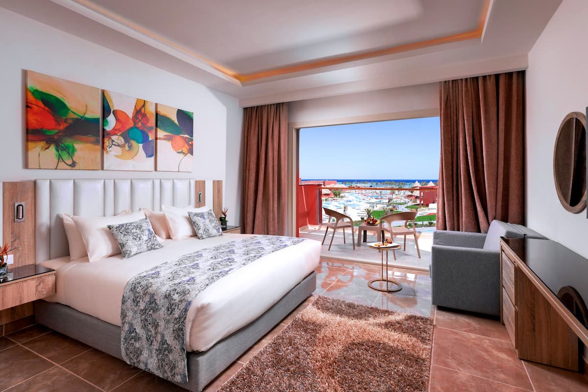 A modern hotel room with a large bed, a balcony with a view of the beach, and artwork on the wall.