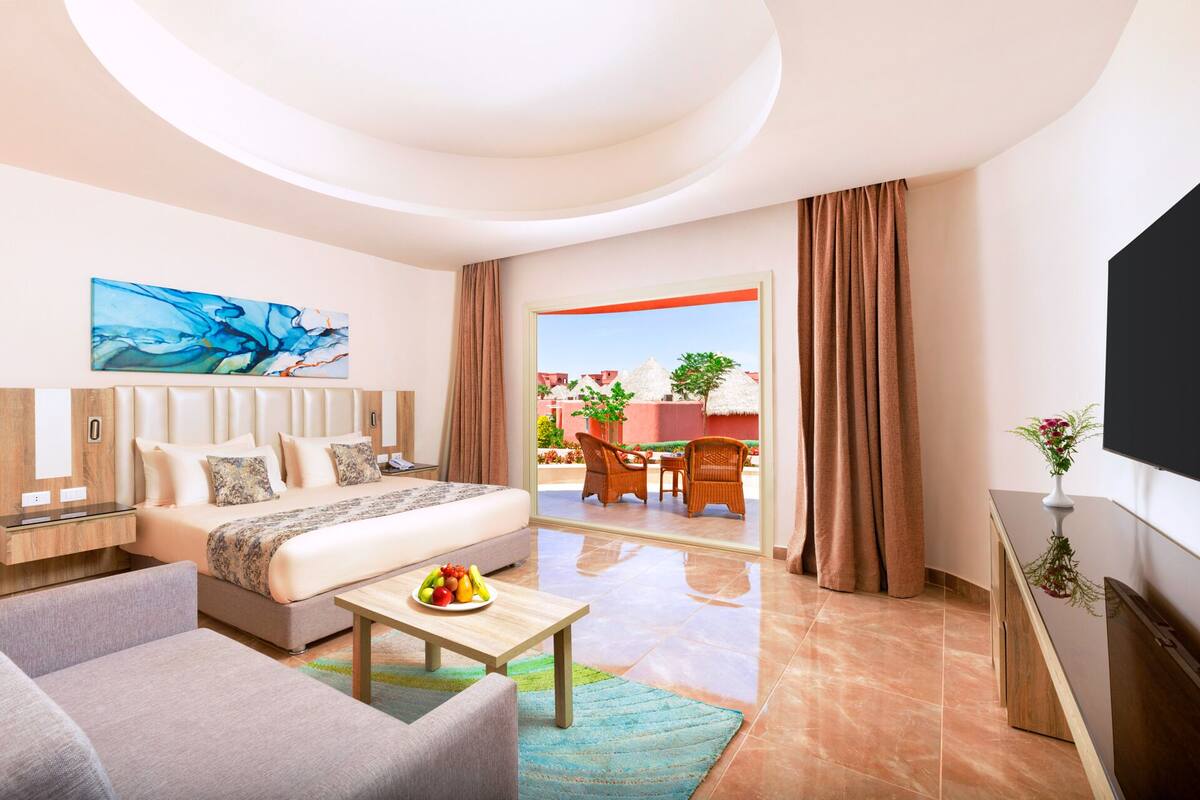 A modern hotel room with a bed, a sofa, a coffee table with fruit, a TV, and a balcony with outdoor seating.