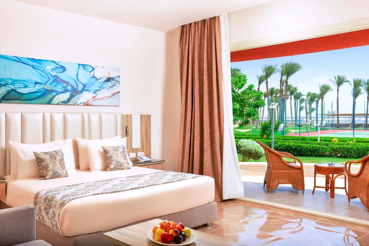 A hotel room with a bed, a desk, and a view of a landscaped garden with palm trees.