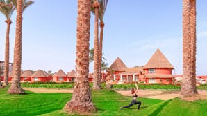 Un complexe hôtelier doté de bâtiments aux toits de chaume, de palmiers et où une personne pratique une posture de yoga.
