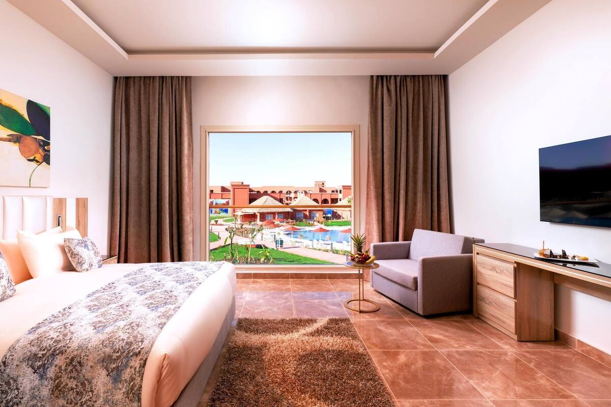 A hotel room with a bed, a desk, a chair, a TV, and a view of a pool and buildings.